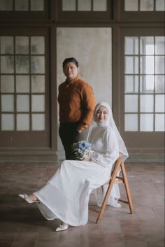 a man standing next to a woman in a white dress on a chair with a veil over her head