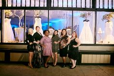 four women standing in front of a window with wedding gowns on display behind them