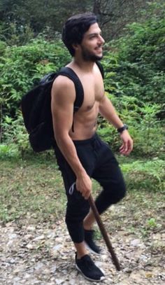 a man with no shirt on is holding a stick and walking in the woods while wearing all black