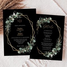 two black and gold wedding cards with greenery in the middle, on top of a white blanket