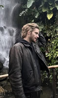 a man standing in front of a waterfall with his hand on his head and wearing a leather jacket