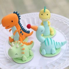 two small toy dinosaurs sitting on top of a white cake plate, one with an orange and the other green