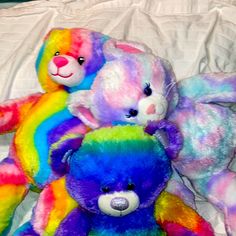 three rainbow colored teddy bears sitting next to each other on a white sheeted bed