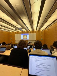 several people sitting at tables with laptops in front of them and a projector screen behind them