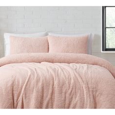 a bed with pink comforter and pillows in front of a brick wall that has a window