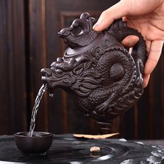 a person is pouring water into a dragon shaped teapot on top of a table