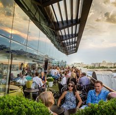 many people are sitting at tables outside near the water and some have drinks in their hands