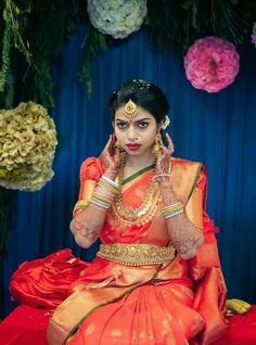 a woman in an orange and gold sari sitting on a red cloth with flowers behind her