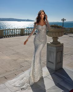 a woman in a wedding dress standing on a sidewalk near the water with her hand up to her face