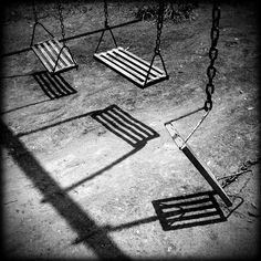 black and white photograph of swings in an empty park with shadows on the concrete ground