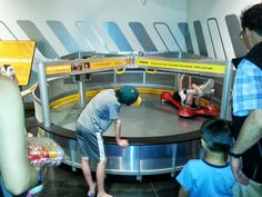 several people standing around in front of a round metal table with two children on it