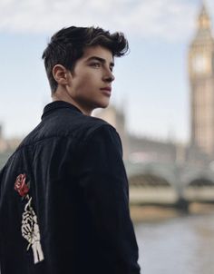 a young man standing in front of a river with a clock tower in the background