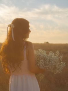 a woman with long hair standing in the middle of a field holding a white flower