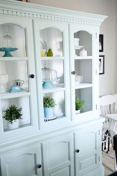 a white china cabinet with glass doors and plants on it's shelves in a dining room