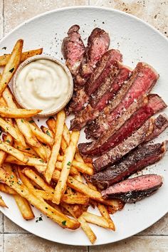 steak and french fries on a white plate with ranch dressing next to it, sitting on a tiled surface