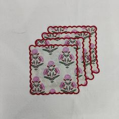 three embroidered napkins with pink flowers on white paper and red trim around the edges