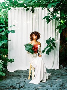 a woman sitting on a chair holding flowers in front of a white curtain with greenery