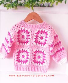 a pink and white crocheted sweater hanging on a wooden hanger next to a plant