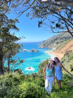 two women standing on top of a lush green hillside next to the ocean and trees