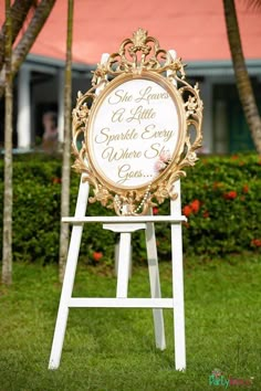 a white and gold sign sitting on top of a lush green field next to palm trees