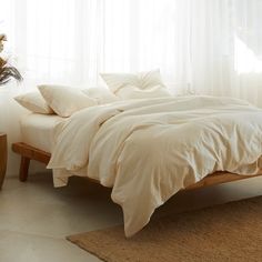 a bed with white sheets and pillows in a room next to a plant on the floor