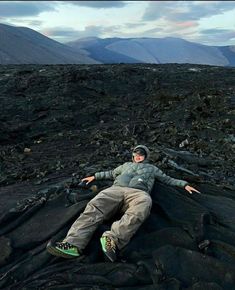 a man laying on top of a black rock covered ground with mountains in the background