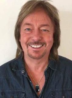 a smiling man with long hair wearing a blue button up shirt and gold chain necklace