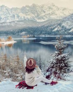 a woman sitting on top of a snow covered hill next to a lake with mountains in the background