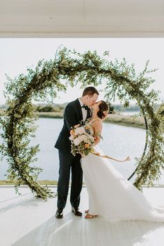 a bride and groom kissing in front of a circular floral arch with greenery on it