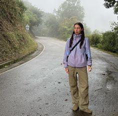 a woman with long hair standing on the side of a road in front of trees