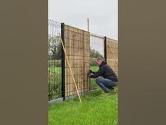 a man leaning against a fence to pick up something from the grass in front of him