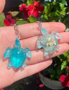 a hand holding two sea turtle charms in it's palm, with flowers in the background