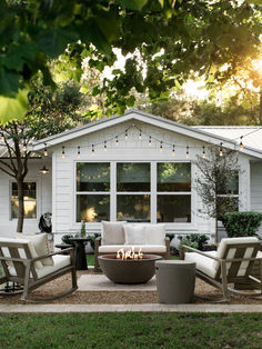 an outdoor fire pit surrounded by chairs and tables in front of a white house with string lights