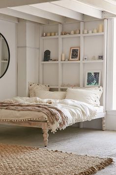 a white bed sitting under a window next to a shelf filled with pillows and blankets