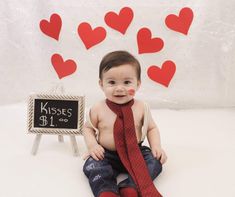 a baby sitting on the floor wearing a red tie and holding a sign with hearts