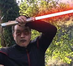 a man holding a light saber over his head with trees in the back ground behind him