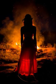 a woman standing in front of a fire with her back to the camera, wearing a long red dress