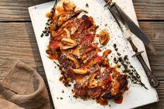 a white plate topped with meat covered in sauce next to a pair of knives and fork