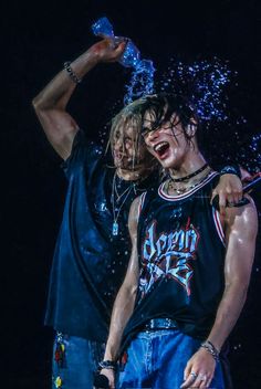 two young men standing next to each other with their arms around one another and water pouring over them