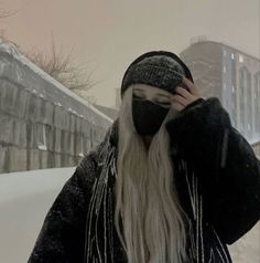 a man with long white hair wearing a black hat and coat in the snow while covering his face