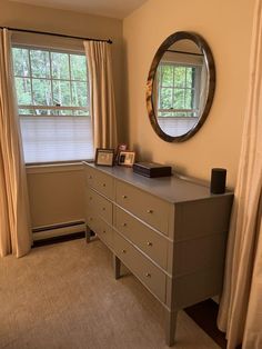 a bedroom with a dresser, mirror and window in the corner next to curtains on the windowsill