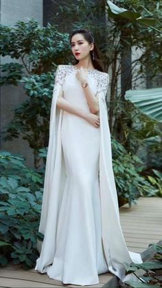 a woman in a white gown and cape standing on a wooden deck surrounded by greenery