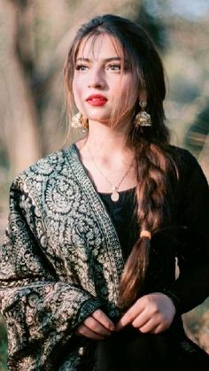 a woman with long hair wearing a black dress and gold earrings, standing in the woods