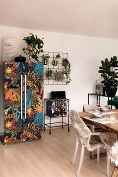 a dining room table with chairs and a refrigerator in the corner, next to potted plants