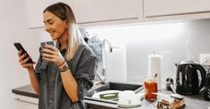 a woman standing in a kitchen holding a cell phone and looking at the screen while smiling