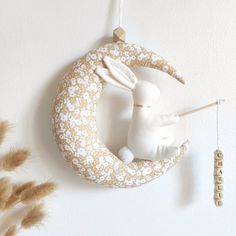 a white rabbit sitting on top of a crescent shaped decoration hanging from a wall next to dry grass