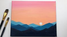 an acrylic painting of mountains with the sun setting in the distance and two paintbrushes next to it