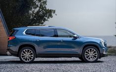 a blue chevrolet suv parked in front of a house next to the ocean and trees