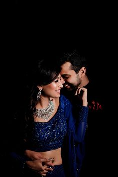 a man and woman standing next to each other in front of a dark background with their arms around each other