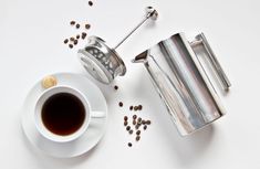 a cup of coffee next to a grinder and beans on a white table top